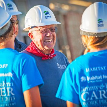 A group of people in hard hats and blue shirts converse, smiling, at a Habitat for Humanity work site.