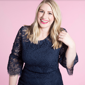 Woman smiling, wearing a blue lace dress, standing against a pink background.