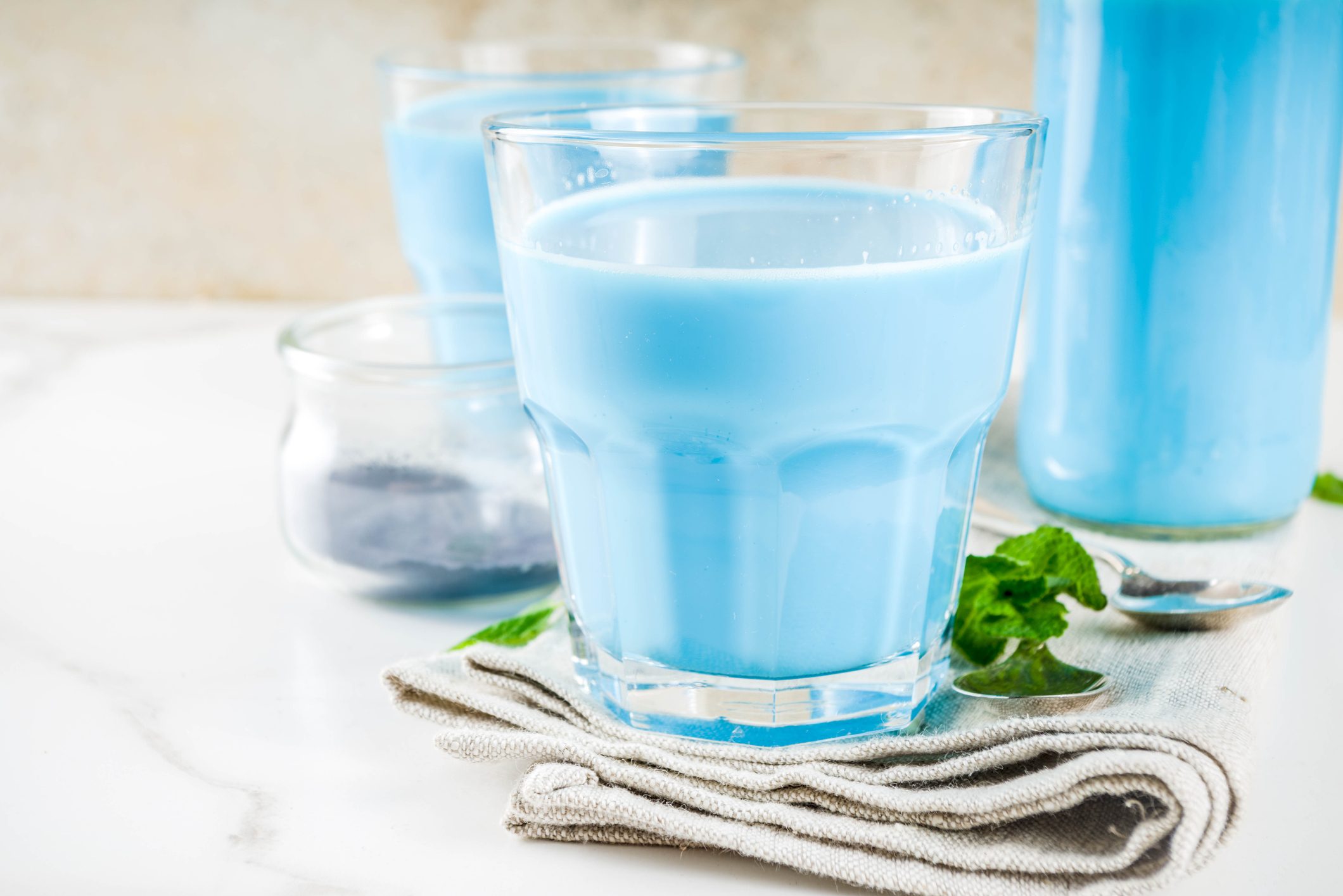 Blue milk fills a glass, resting on a folded napkin, surrounded by mint leaves and additional glasses on a white surface.