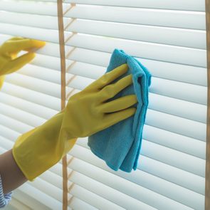 Gloved hands clean window blinds with a blue cloth in a brightly lit room.