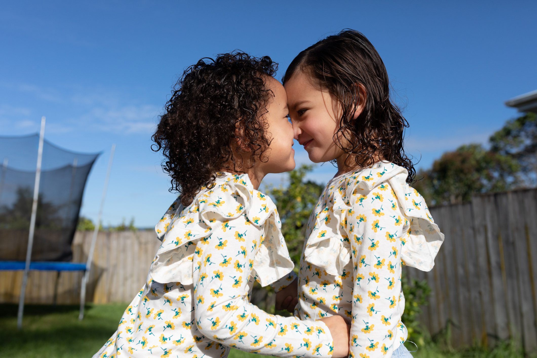 Two children wearing matching floral tops touch noses, smiling affectionately in a sunny backyard with a trampoline and wooden fence.