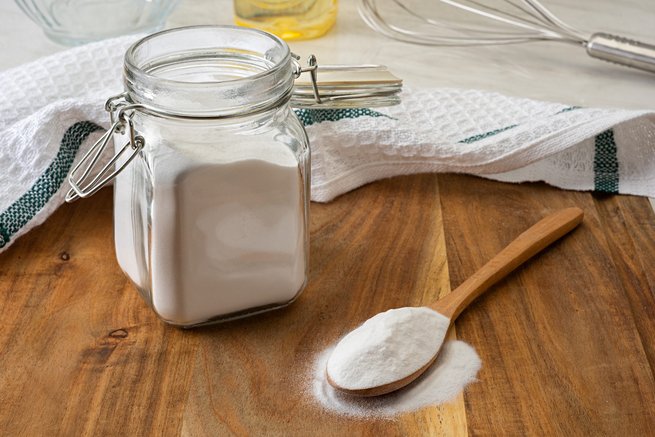 A jar filled with white powder stands next to a wooden spoon on a wooden surface, surrounded by a towel and whisk.