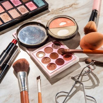 Makeup products are scattered on a marble surface, featuring eyeshadow palettes, brushes, lipstick, an eyelash curler, and a blending sponge.