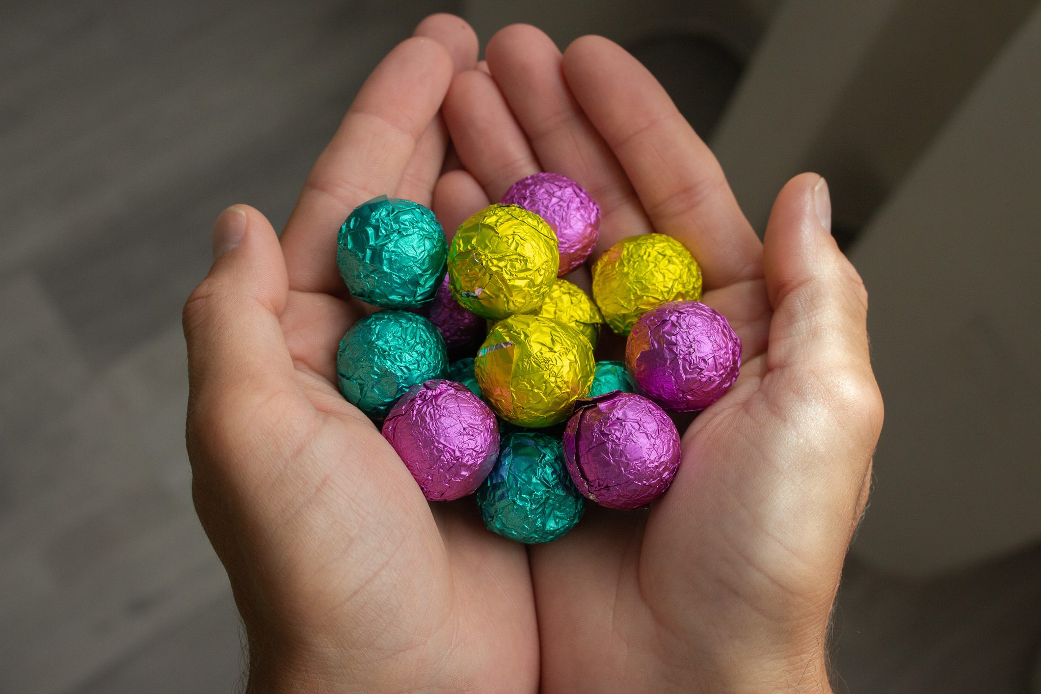 Hands hold colorful foil-wrapped chocolate balls, arranged in cupped palms, against a softly blurred, neutral background.
