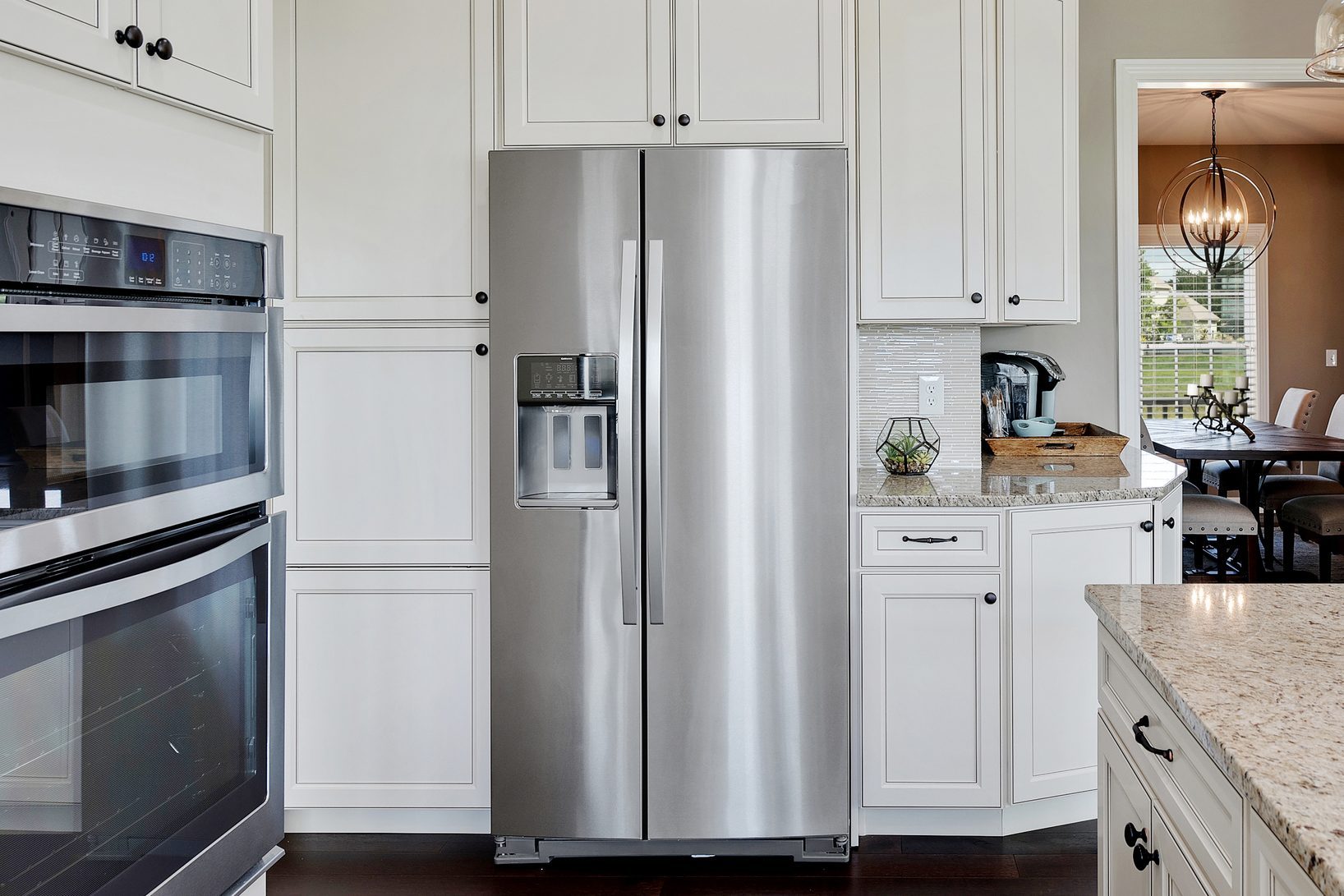 A stainless steel refrigerator stands in a modern kitchen, flanked by white cabinets, an oven, and a view of a dining room.