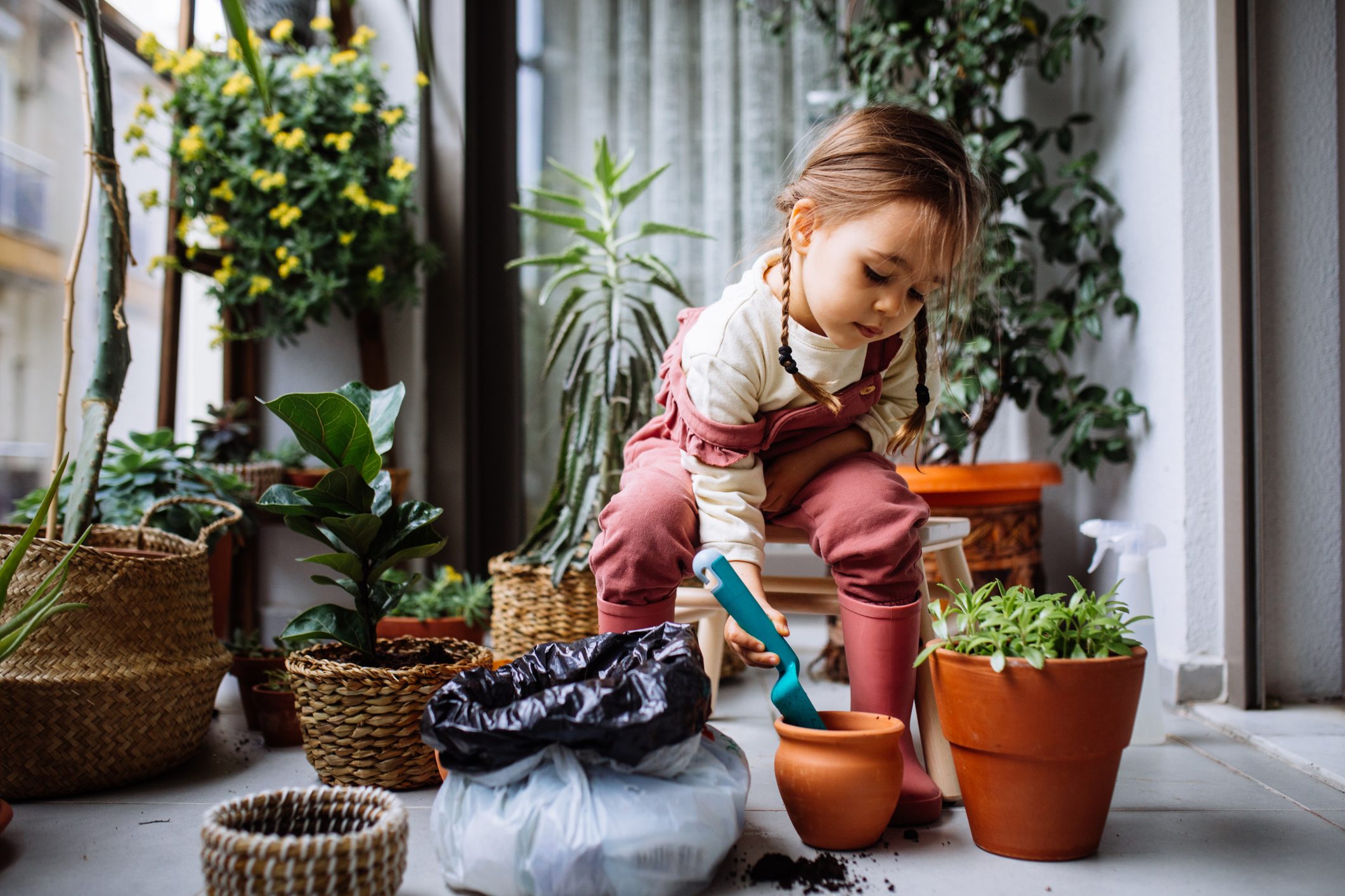 Child gardening indoors, using a small blue trowel to plant in a clay pot, surrounded by various potted plants.