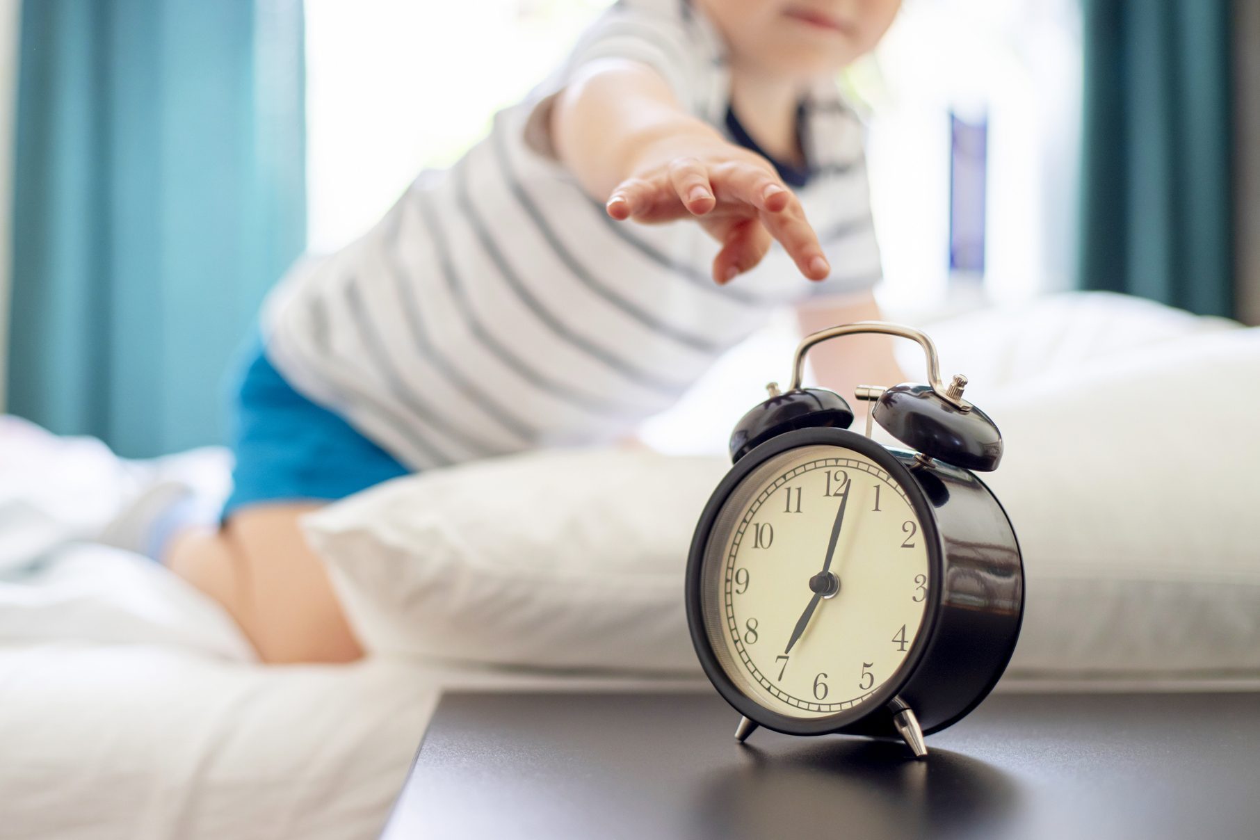 Alarm clock sits on bedside table; child reaches towards it, partially lying on a bed with white pillows and blue curtains in the background.