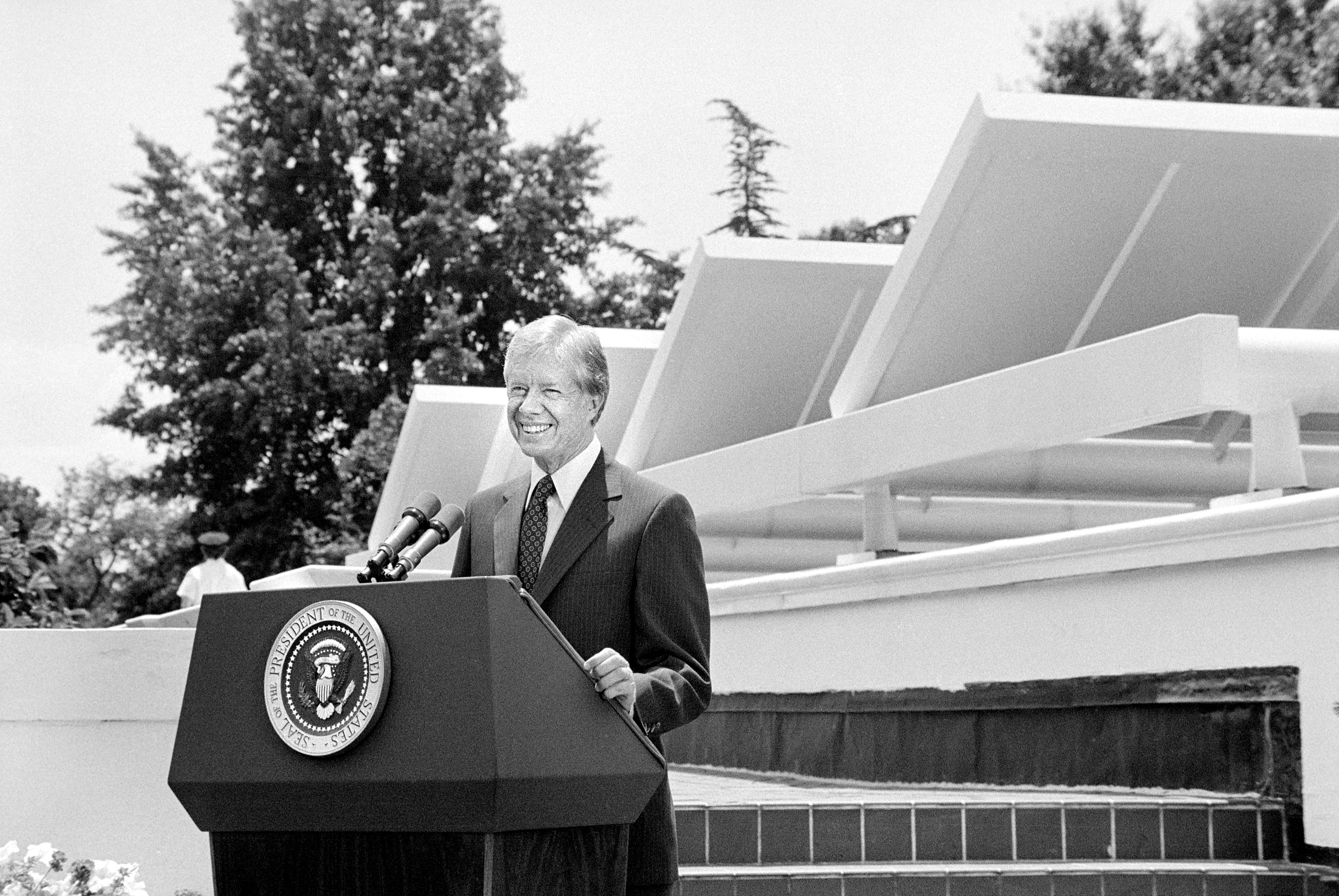 A man speaks at a podium with microphones, surrounded by trees, near large solar panels and a seal of the President of the United States.