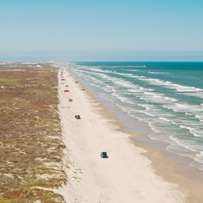 Vehicles drive along a sandy beach, bordered by ocean waves on one side and grassy dunes on the other, under a clear blue sky.
