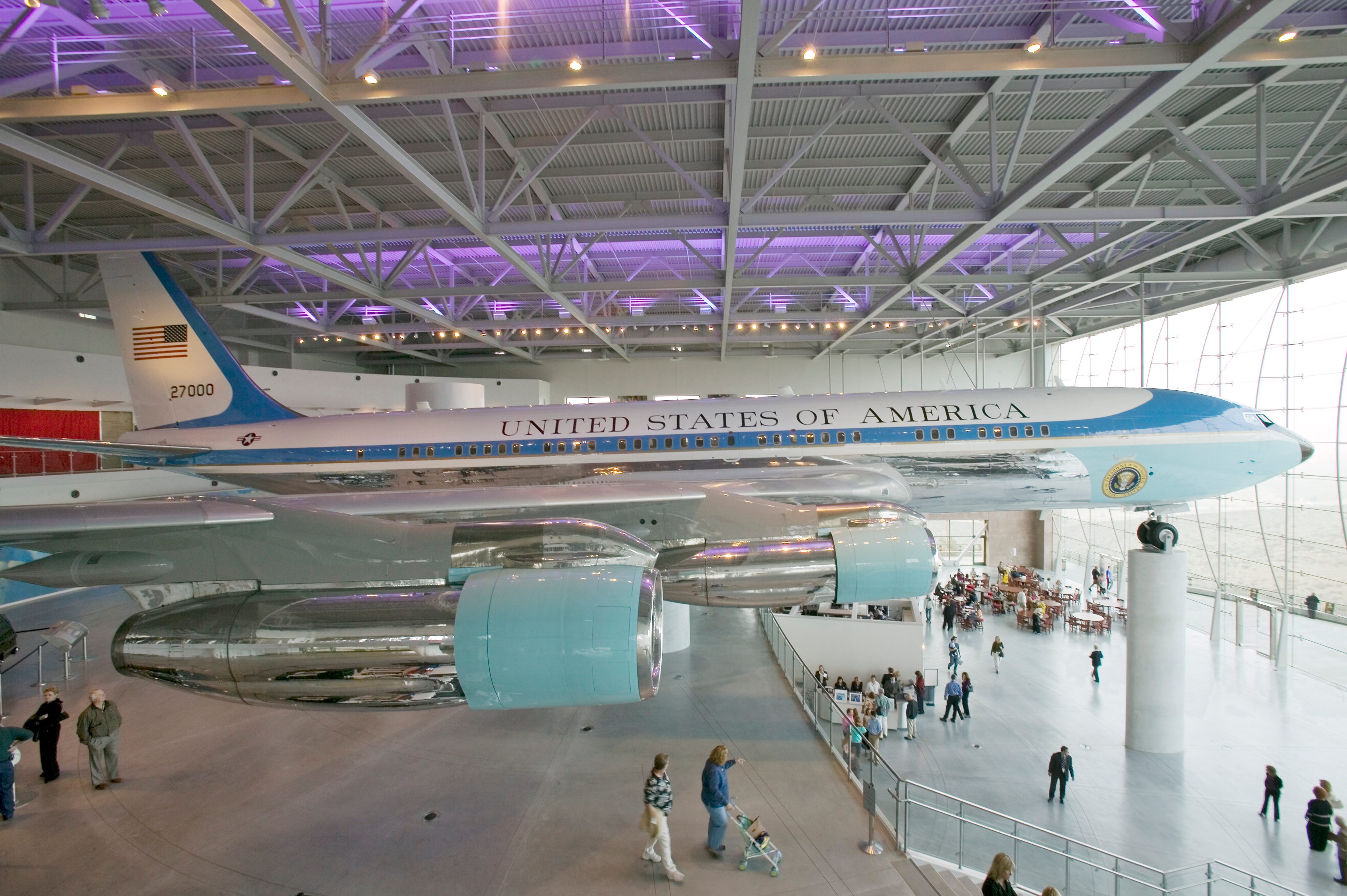An airplane displayed indoors, labeled "UNITED STATES OF AMERICA," hovers above visitors walking through a spacious, modern exhibition hall.