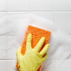 A yellow-gloved hand scrubs a soapy white tiled wall with an orange sponge, creating foam.