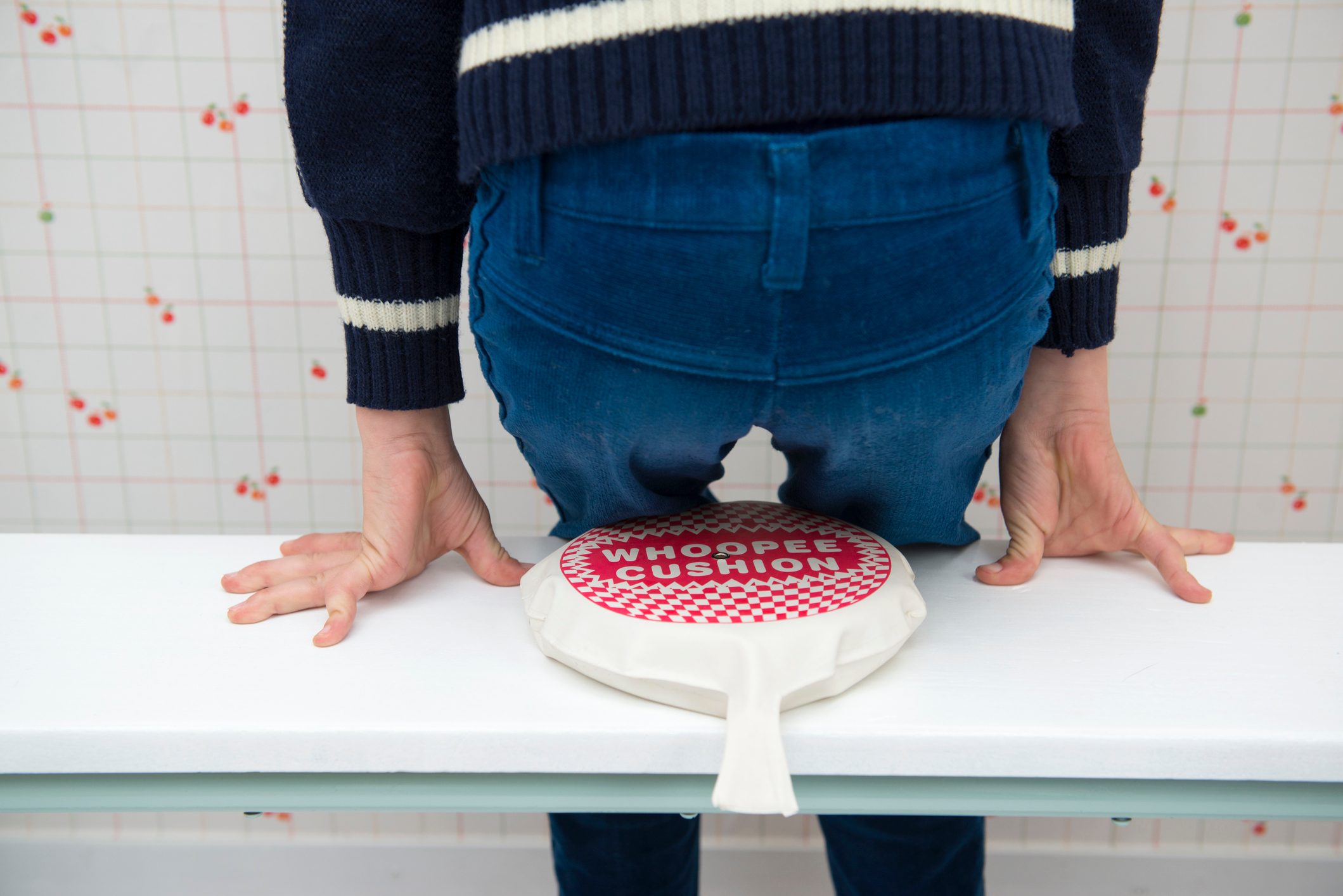 Whoopee cushion placed under a person sitting on a white bench; background with cherry-patterned wallpaper.
