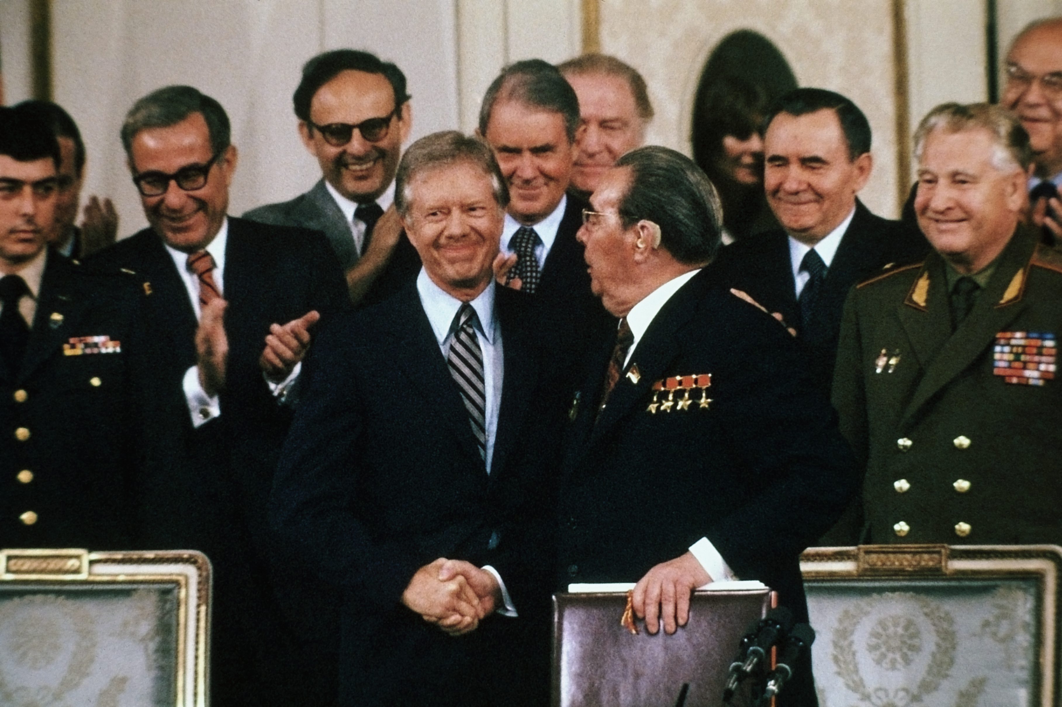 Two men shake hands, surrounded by applauding officials, in a formal setting with microphones and ornate chairs in the foreground.