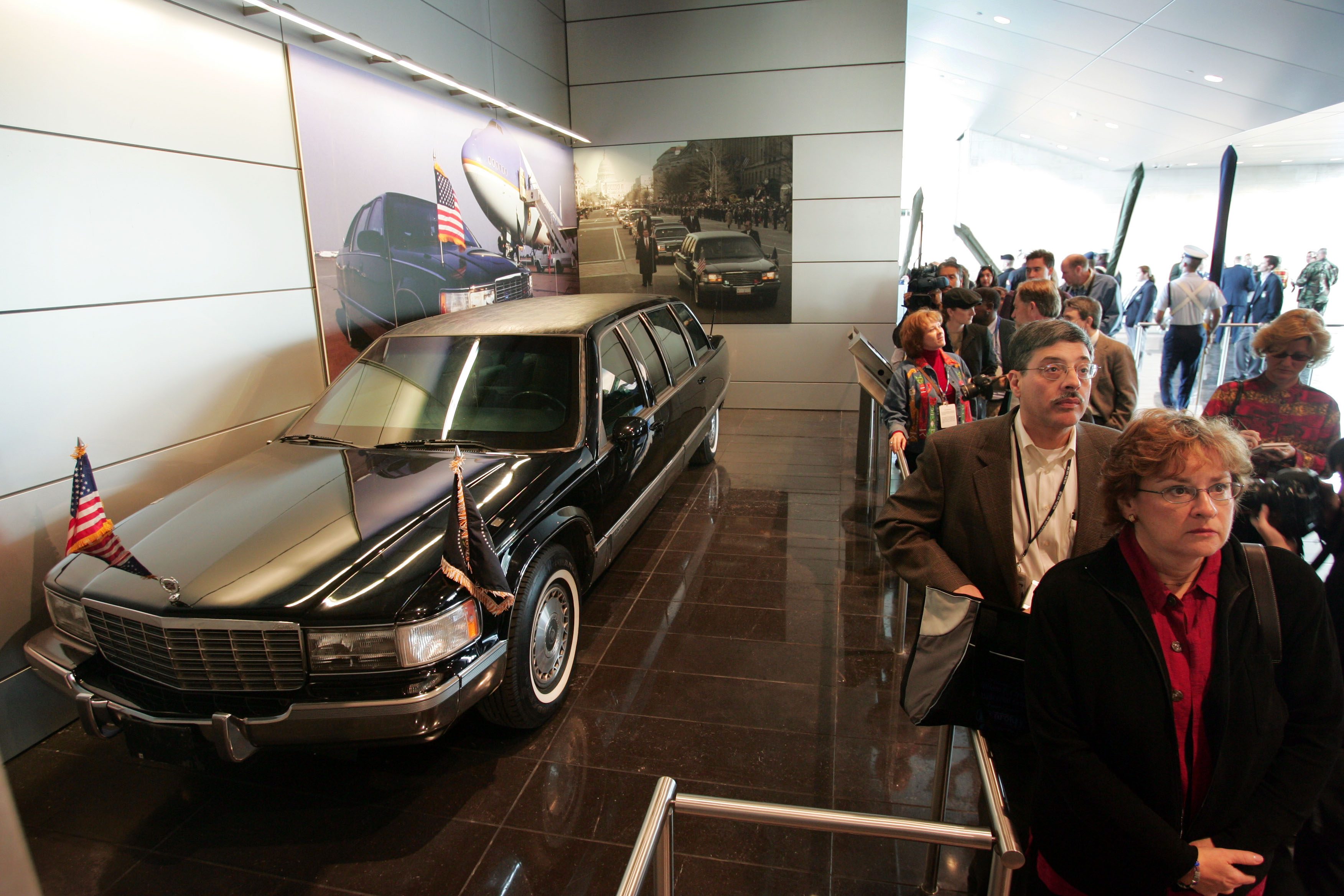 A black limousine is displayed indoors, with American flags on the hood. People line up to view it in a museum-like setting.