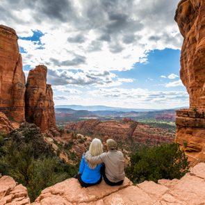 Two people sit together, embracing, on a cliff edge overlooking a vast, scenic desert canyon landscape under a partly cloudy sky.