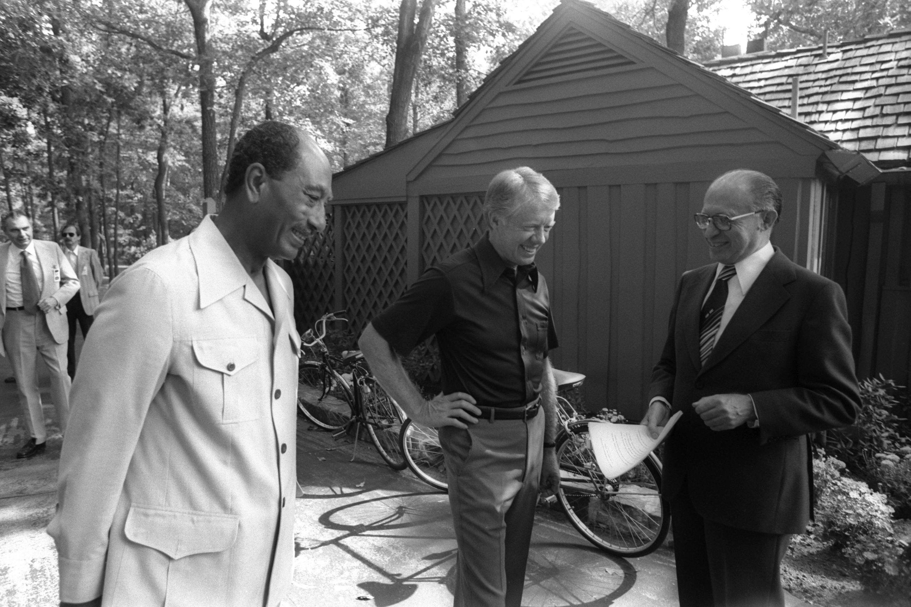 Three men in discussion, smiling, outdoors near wooden structures and bicycles in a wooded setting. One holds papers, appearing engaged and relaxed.