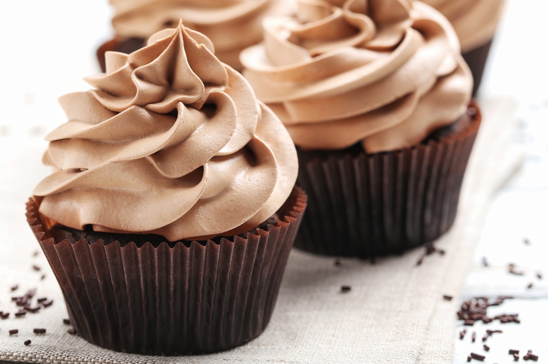 Cupcakes topped with swirled chocolate frosting sit on a light fabric, surrounded by scattered chocolate sprinkles.