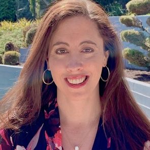 A person smiles outdoors, wearing hoop earrings and a floral top. The background features trimmed bushes and a blue-painted retaining wall.