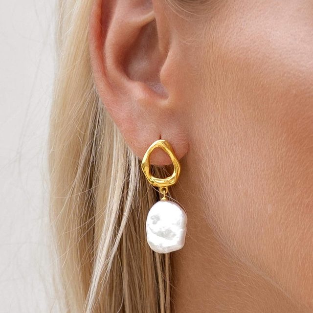 Earring dangling from ear, featuring a gold loop and large irregular white pearl, against light-colored hair and skin.