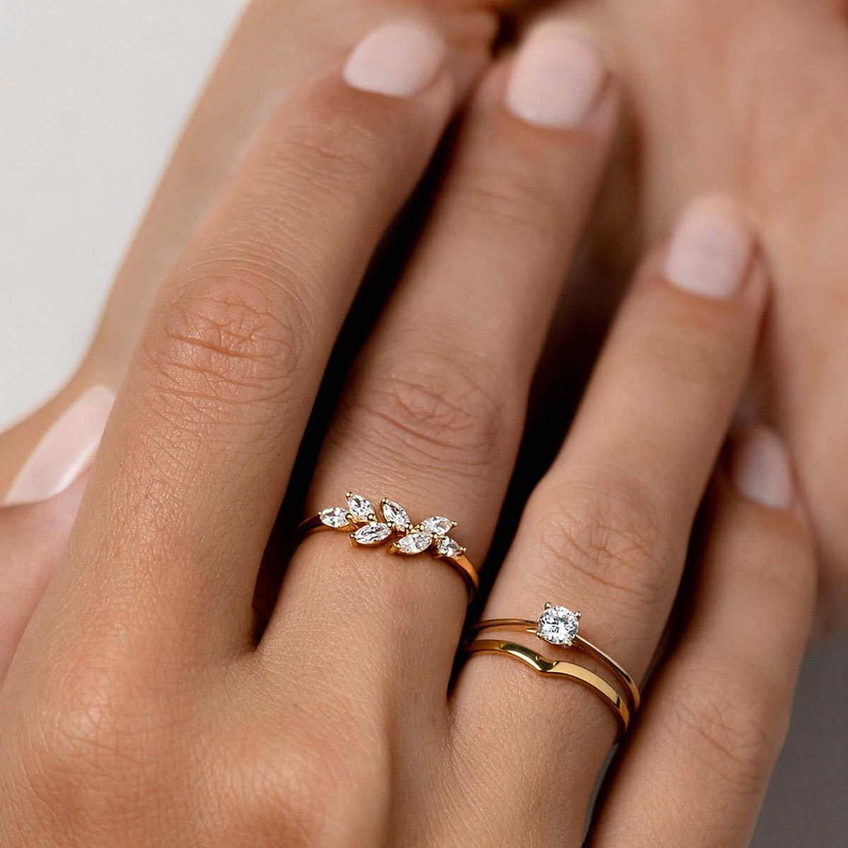 Hands display two gold rings; one features leaf-shaped stones, the other has a single round gemstone. The background is a neutral surface.