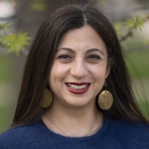 A woman smiles, wearing large round earrings, in an outdoor setting with green leaves blurred in the background.