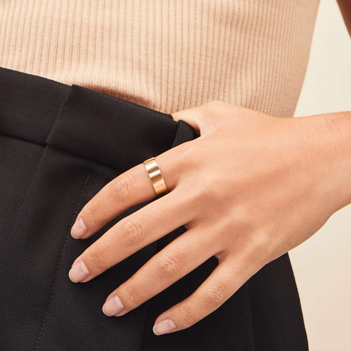 A hand with a gold ring rests on black pants, with a beige ribbed top in the background.