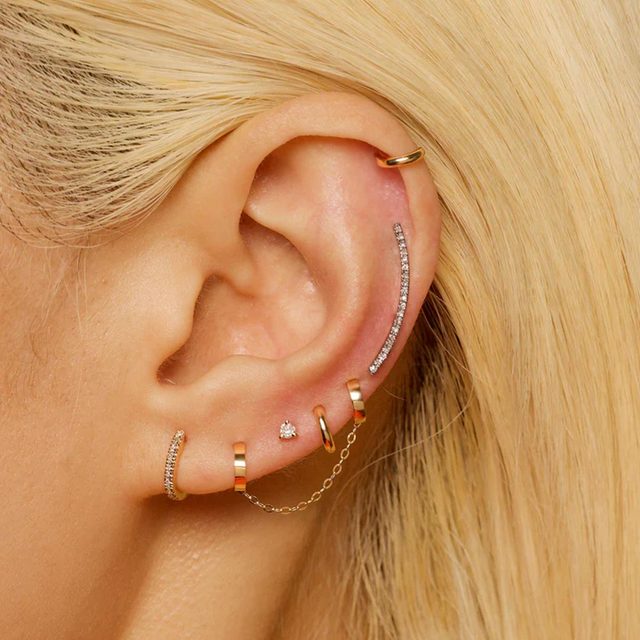 Earrings adorn an ear; multiple gold hoops and studs embellished with diamonds, some linked by a chain, amidst blonde hair.