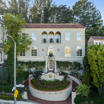 Large, elegant house stands in a lush, hilly neighborhood, surrounded by trees and manicured shrubs; a "Dead End" sign marks the corner.