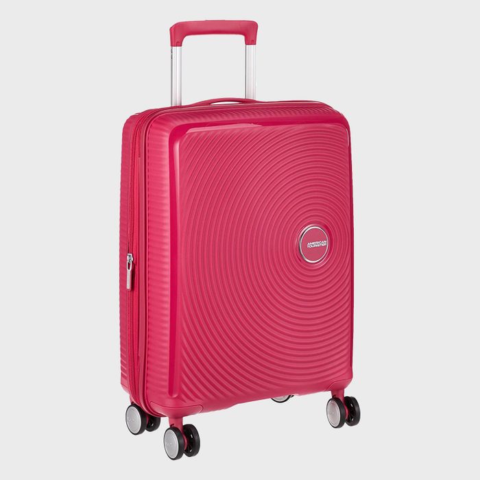 Red suitcase stands upright with four wheels, featuring a textured circular pattern, retractable handle, and American Tourister logo, against a light gray background.