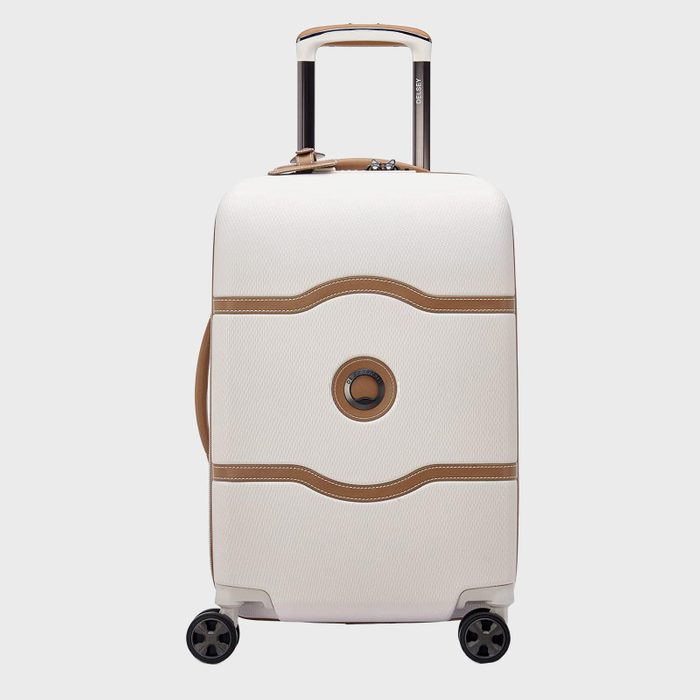 A white and tan suitcase stands upright with an extended handle, featuring four wheels, against a plain background.
