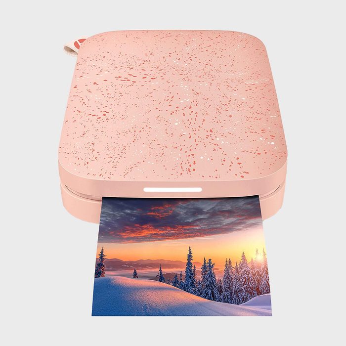 A pink portable printer is ejecting a vibrant landscape photo, featuring snow-covered trees at sunset, against a plain background.