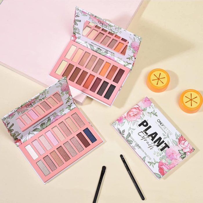 Two open eyeshadow palettes display vibrant shades on a floral background, surrounded by brushes and orange containers with floral box lids.