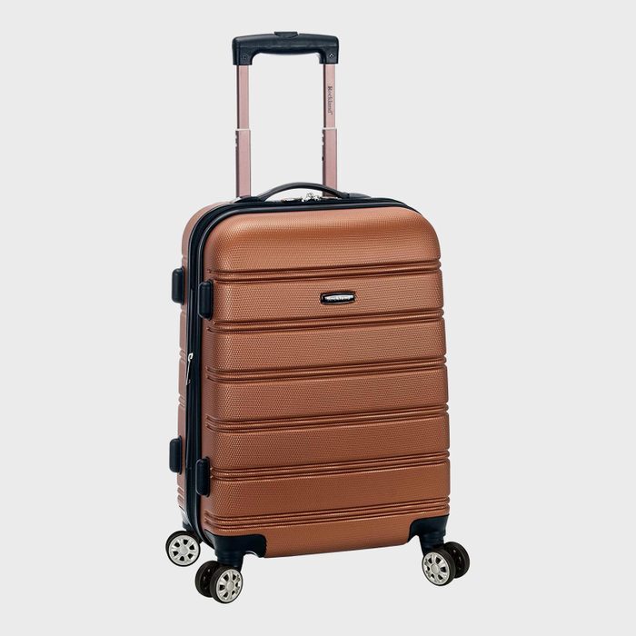 Suitcase stands upright, featuring a textured brown surface and four spinner wheels, with a telescopic handle extended. The background is plain gray.