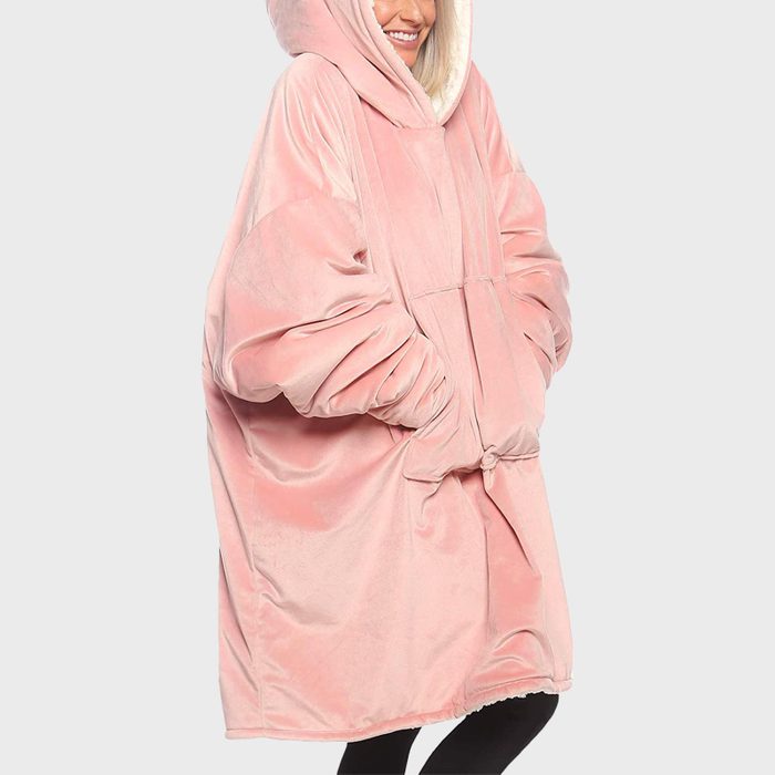 A person wears an oversized, pink, hooded blanket with sleeves, smiling against a plain gray background.