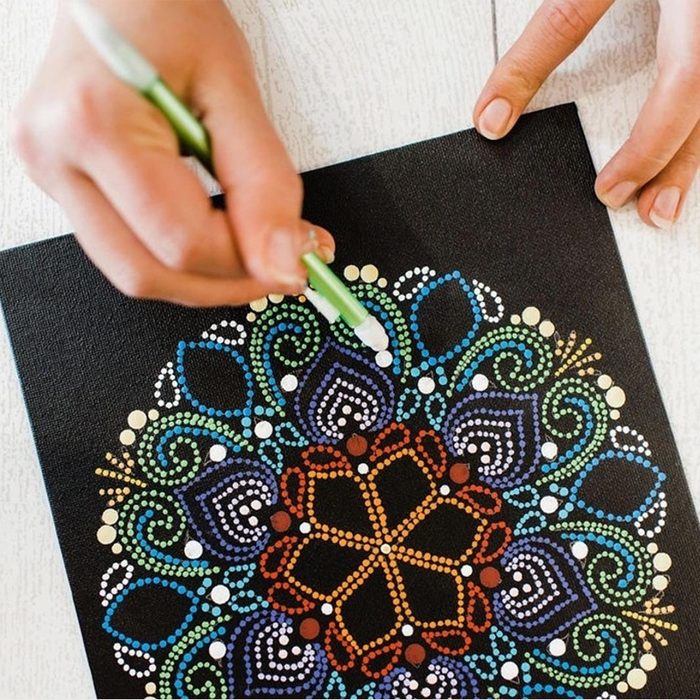 Hand paints colorful dot mandala pattern on black canvas, using a green pen. The artwork features intricate swirling designs and vibrant colors.