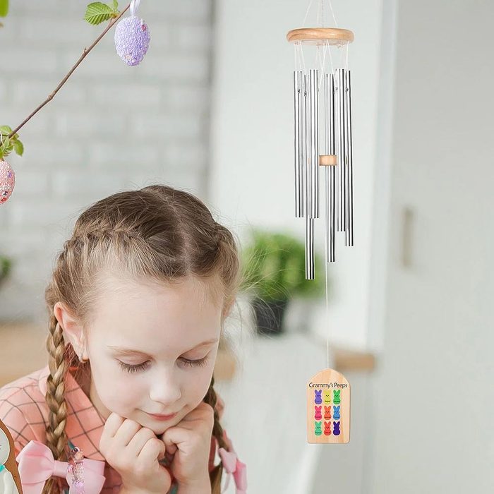 Wind chimes gently sway indoors; a child with braids observes quietly. Nearby branches hold decorative eggs. Text reads: 