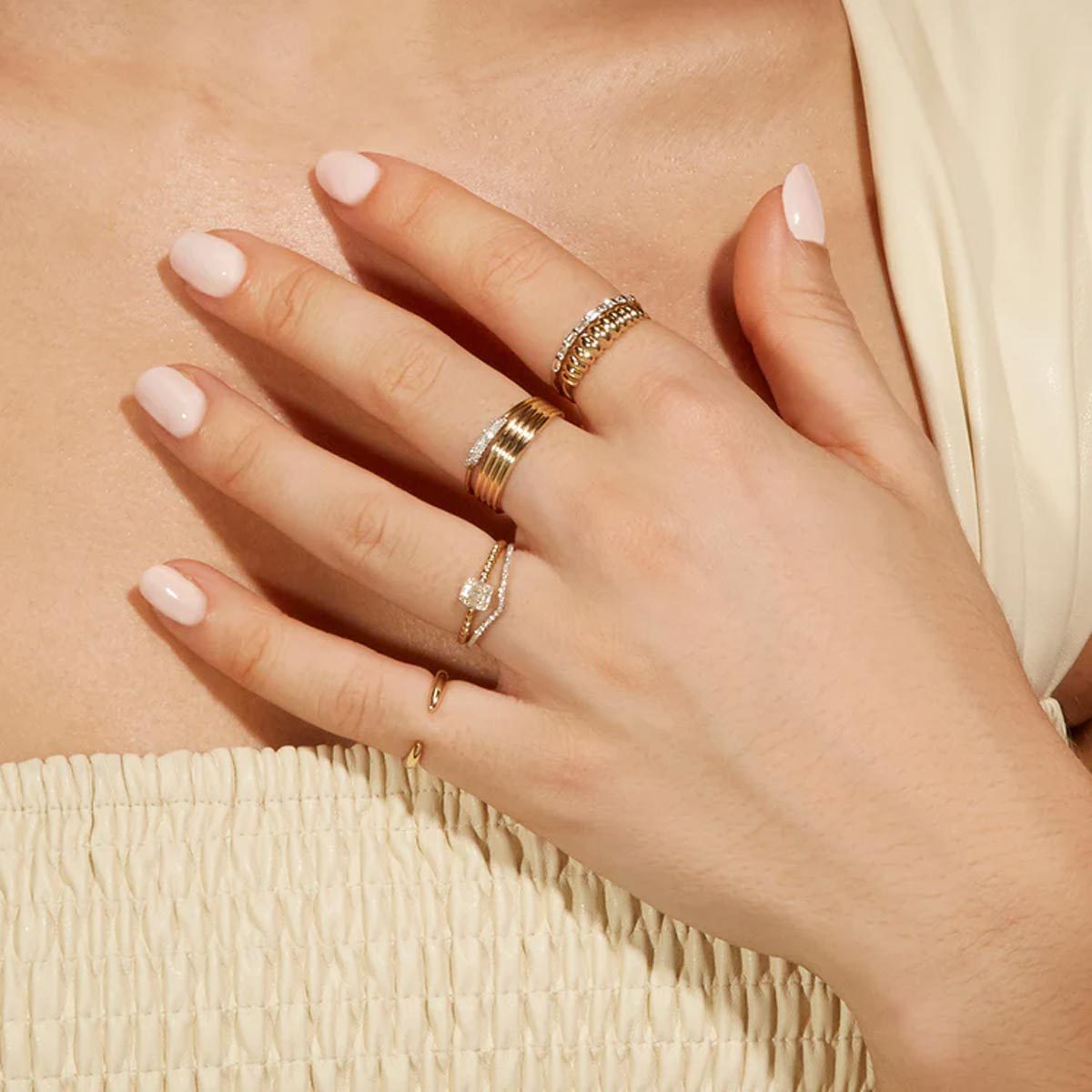 Hand adorned with gold and diamond rings rests gracefully on clothing, featuring pale pink nails and soft, natural lighting.