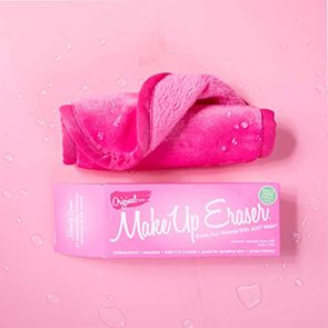 A pink makeup remover cloth, folded and wet, rests on a matching surface beside its branded box, surrounded by water droplets.