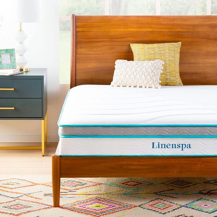 Mattress lies on a wooden bed frame, topped with pillows in a bright room featuring a geometric rug and a modern nightstand with a lamp.