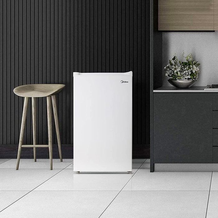A white refrigerator stands beside a wooden stool and a kitchen counter with a plant, set against a dark, striped wall.