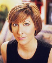 Person sitting, looking towards the camera with a neutral expression, in a cozy indoor environment with soft-focus background elements.