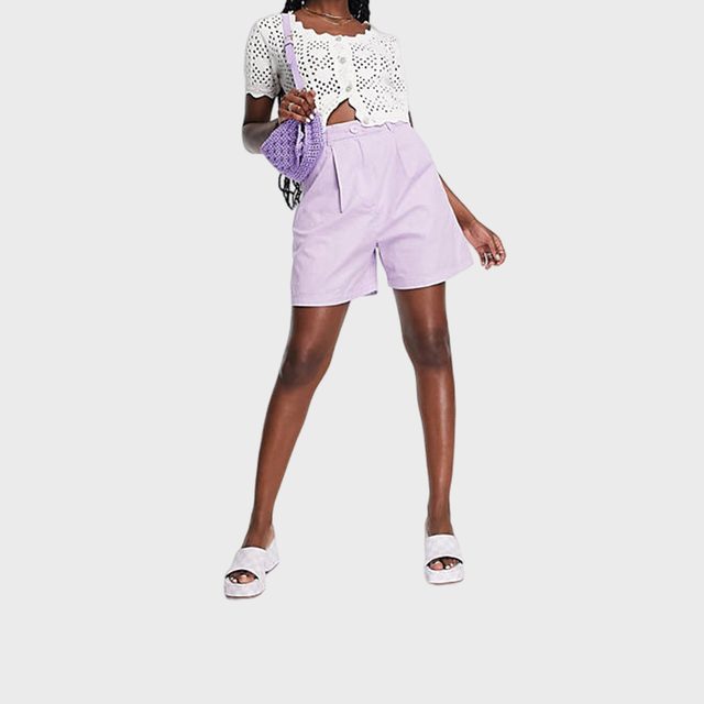 Person standing confidently, wearing a white crocheted top and lilac shorts, holding a purple bag. White sandals complete the casual outfit.