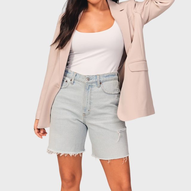 Woman wearing denim shorts, white tank top, and beige blazer, standing with one arm raised against a light gray background.