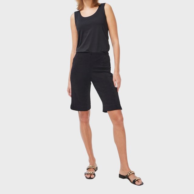Woman wearing black tank top and shorts stands confidently, modeling black sandals with gold chain detail. Plain white background emphasizes her attire.