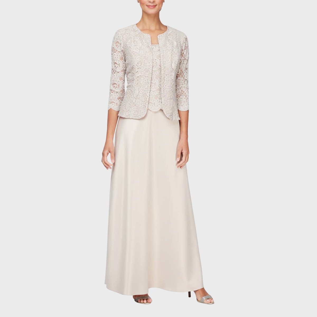 A woman wears an elegant cream lace jacket over a floor-length gown, standing against a plain white background.
