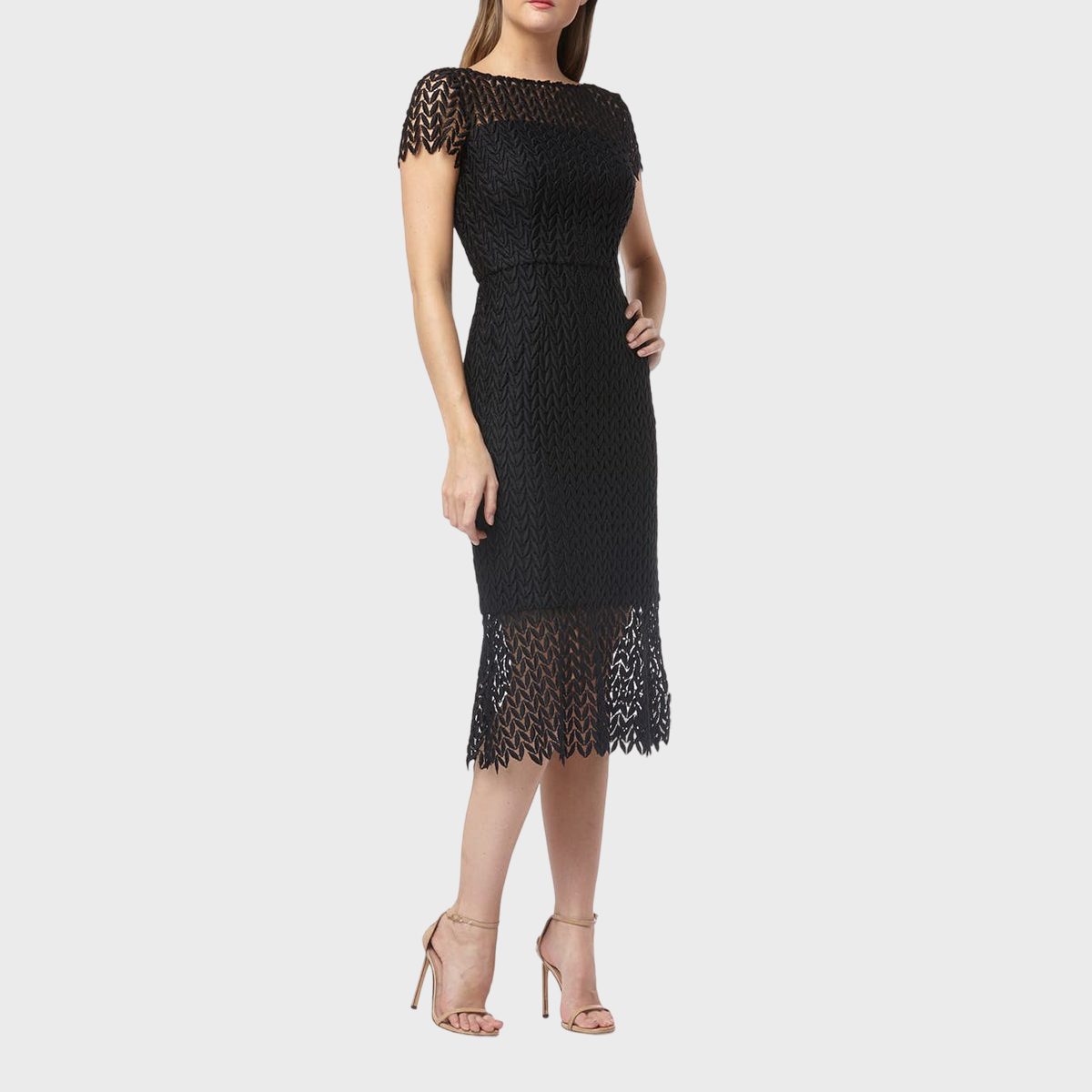 A woman wears a black lace dress, standing in neutral heels against a plain white background.