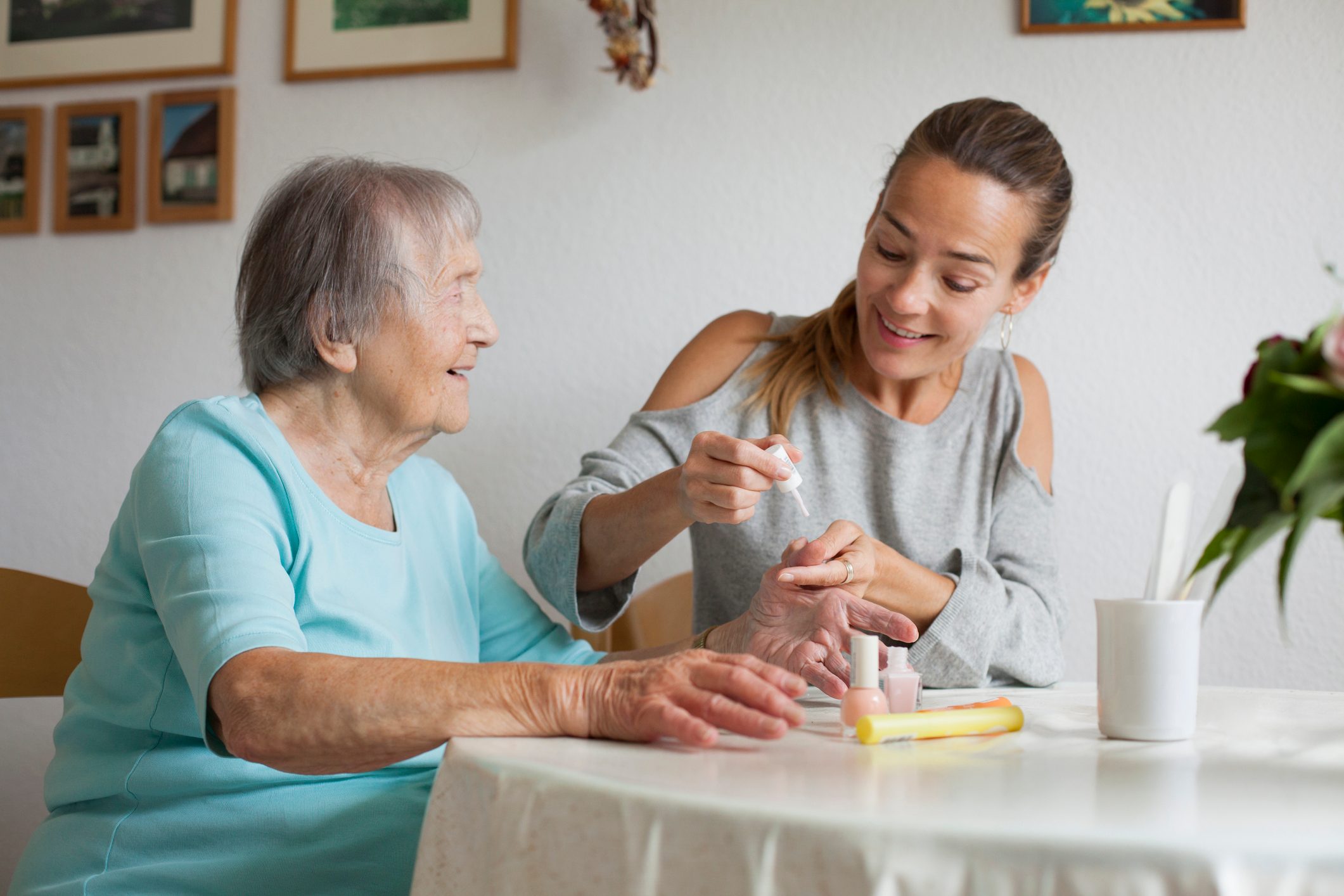 A woman applies nail polish to an elderly woman's nails at a table, with framed pictures on the wall in a cozy setting.