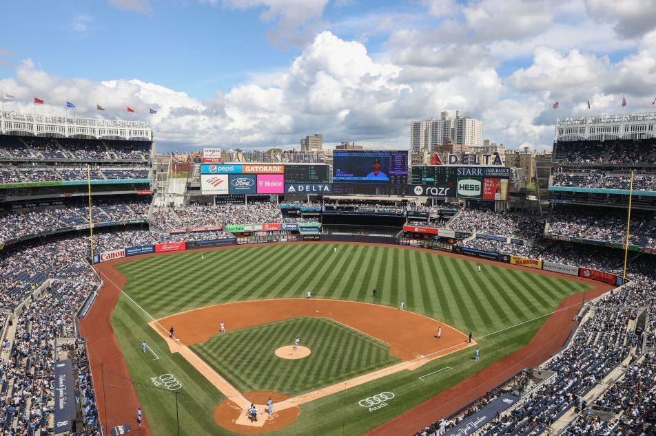 Stadium hosts a baseball game, players on field, surrounded by cheering crowd; blue sky and city skyline visible in background, advertisements line outfield.