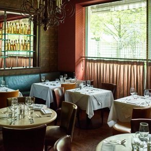 Tables with white cloths holding glassware, set in a dimly lit restaurant with a chandelier, shelves of bottles, and windows with blinds.