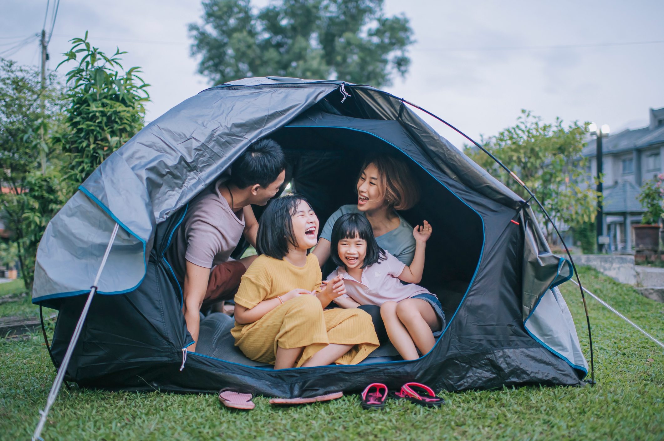 Family sits laughing inside a tent pitched on grass, surrounded by trees and nearby houses, creating a joyful outdoor setting.